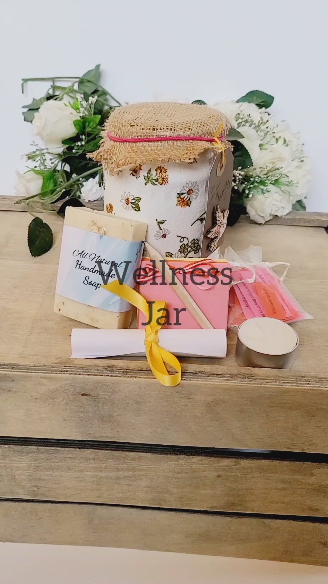 Vdeo of a White jar with bee and flower design, hessian lid with card label with a dove on it. Contents shown outside of the jar, cream soap, small multicoloured scrapbook, short wooden pencil, tealight candle, small organza bag containing coloured paper quotes, white rolled up instruction leaflet tied with yellow ribbon.
