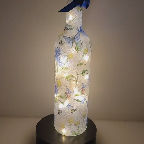 A rotating example of a A lit up wine bottle decorated with blue flowers, blue butterflies, acorns and bumble bee. Cork LED lights inside and blue ribbon around the neck of the bottle