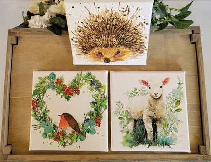 Three nature-themed prints featuring a hedgehog, robin, and lamb on a wooden surface with floral decorations.