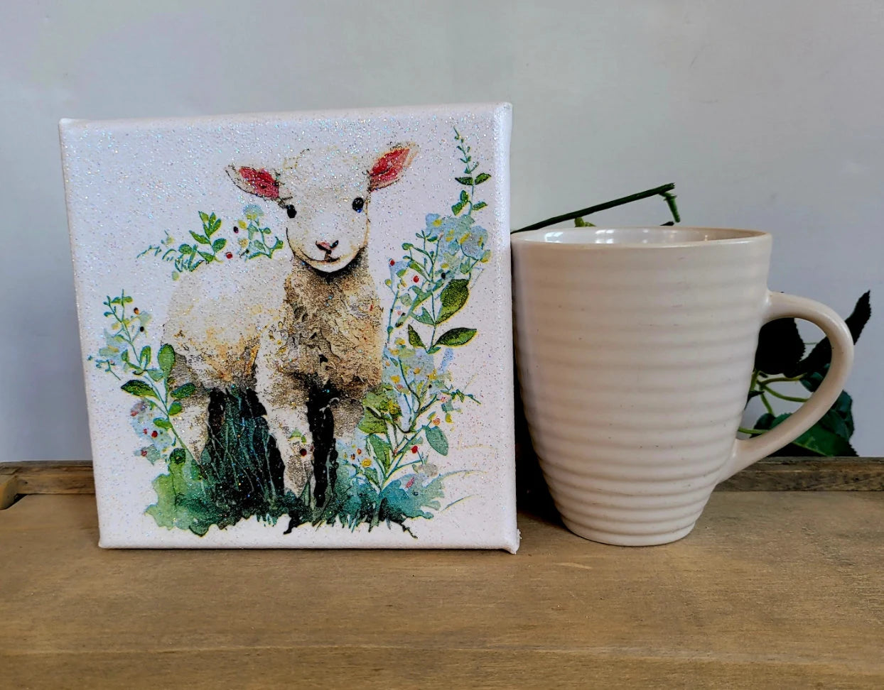 Canvas print of a lamb surrounded by flowers next to a ceramic mug on a wooden surface.
