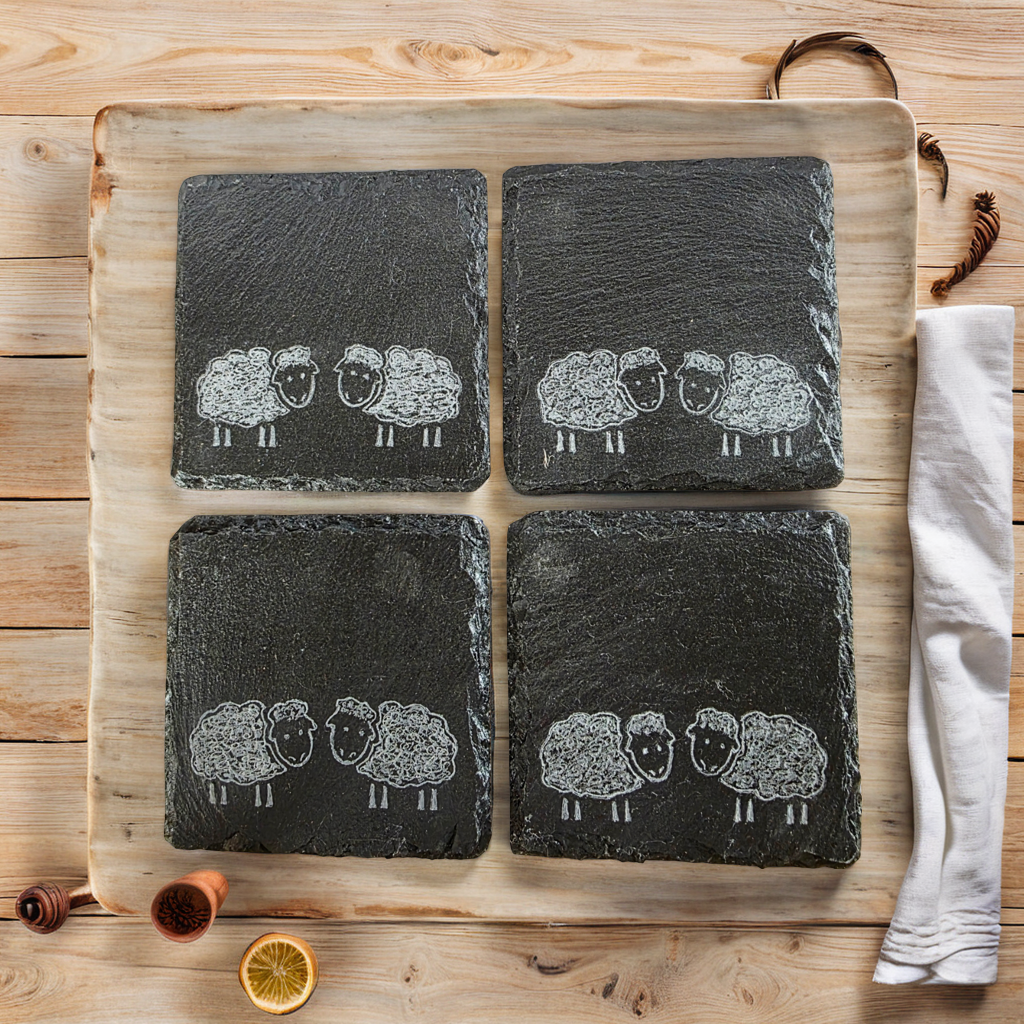 Four slate coasters with sheep illustrations on a wooden board.