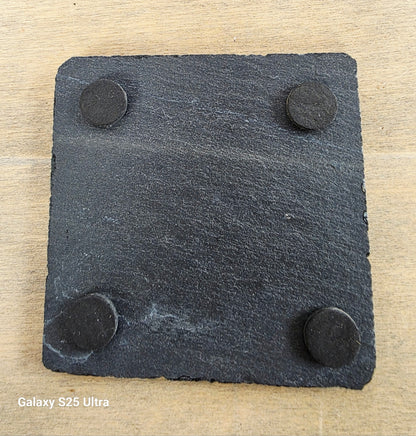 Square slate coaster with four black rubber feet on a wooden surface