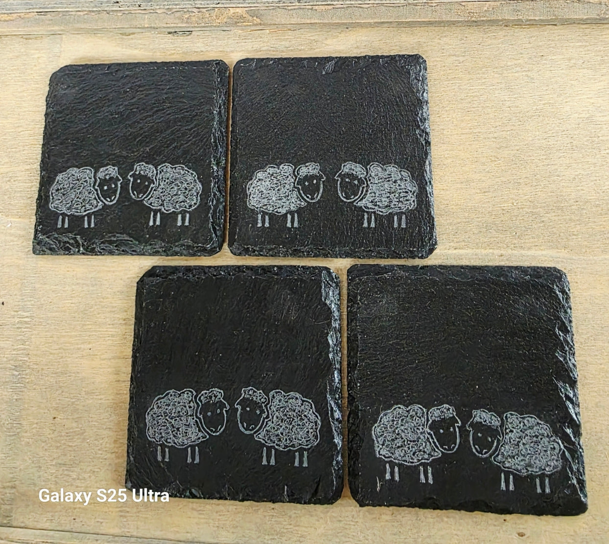 Four black slate coasters with engraved sheep designs on a wooden surface.