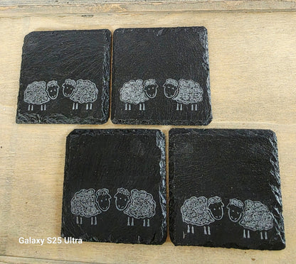 Four black slate coasters with engraved sheep designs on a wooden surface.