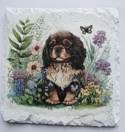 White slate coaster with a painting of a king charles spaniel puppy surrounded by flowers on a white background