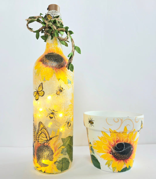 Decorative bottle and pot with sunflower  and bumble bee design, jute twine with leaves around the neck of the bottle. Lit up with cork lights. on a white background