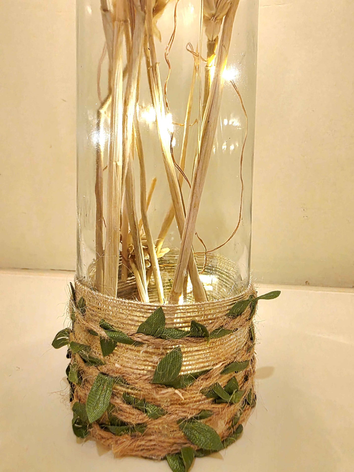 A decorative glass bottle filled with dried stems and warm white fairy lights, wrapped at the base with beige twine and green leaves, set against a plain light background.