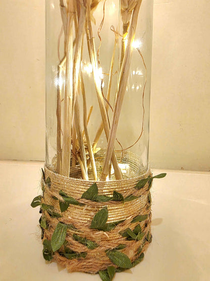 A decorative glass bottle filled with dried stems and warm white fairy lights, wrapped at the base with beige twine and green leaves, set against a plain light background.