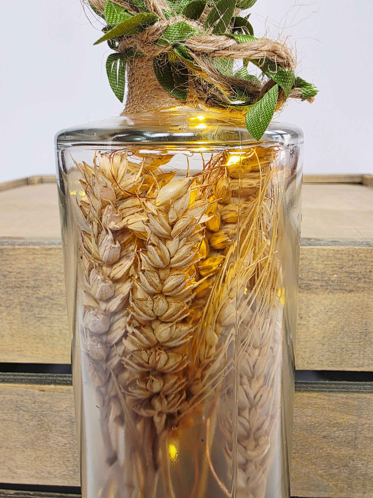 A clear glass vase filled with dried wheat stalks and small warm white fairy lights, topped with a rustic burlap-wrapped neck and green leaves, set against a light wood crate background.