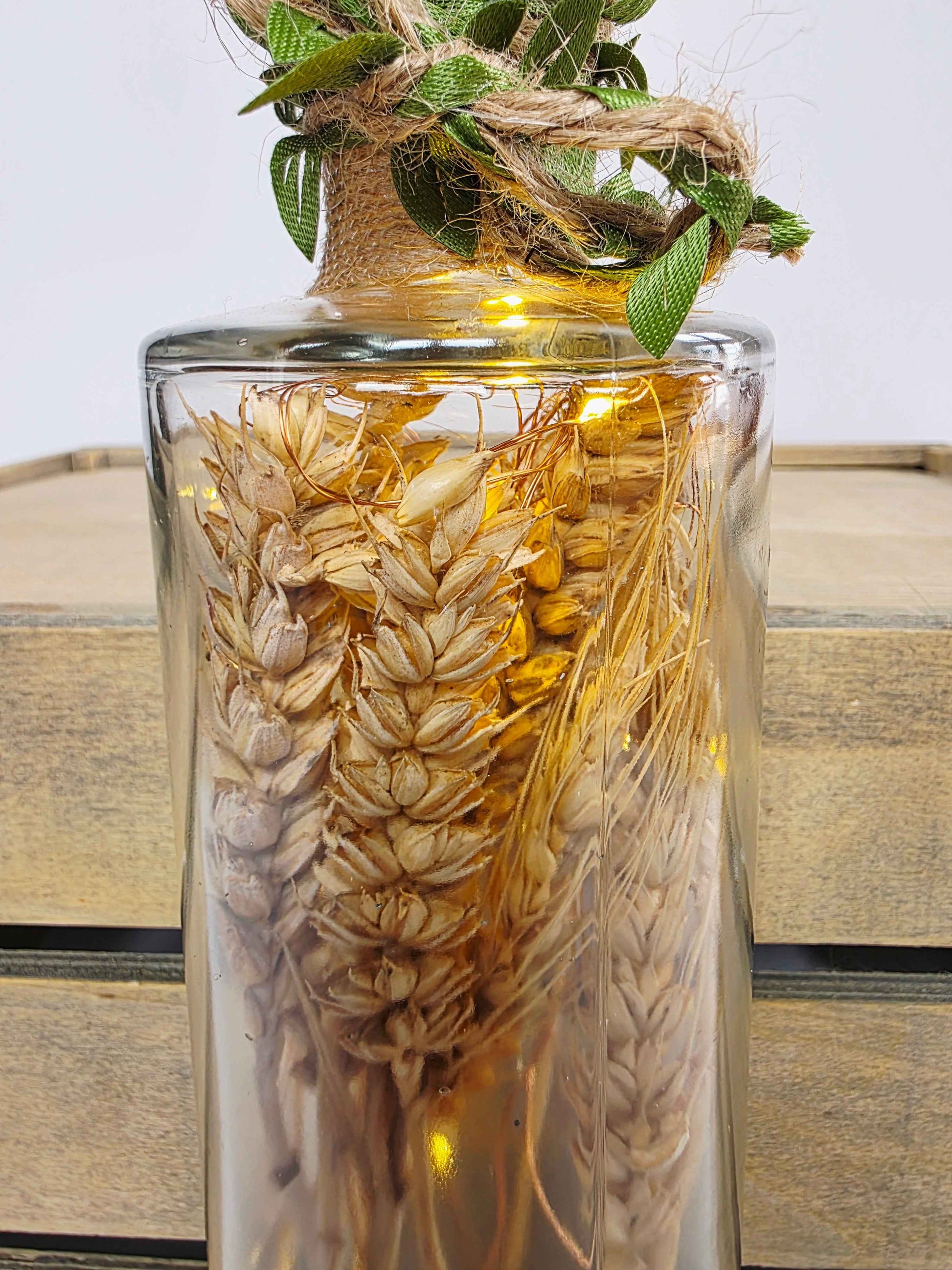 A clear glass vase filled with dried wheat stalks and small warm white fairy lights, topped with a rustic burlap-wrapped neck and green leaves, set against a light wood crate background.