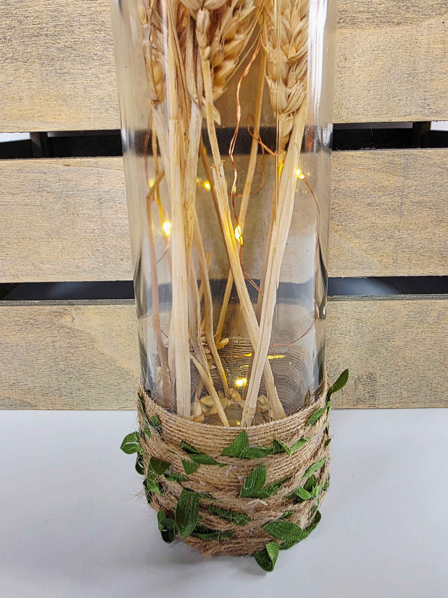 A clear glass bottle with dried wheat stalks and warm white fairy lights, wrapped at the base with natural jute twine and green leaf accents, set against a light wood slat background. 