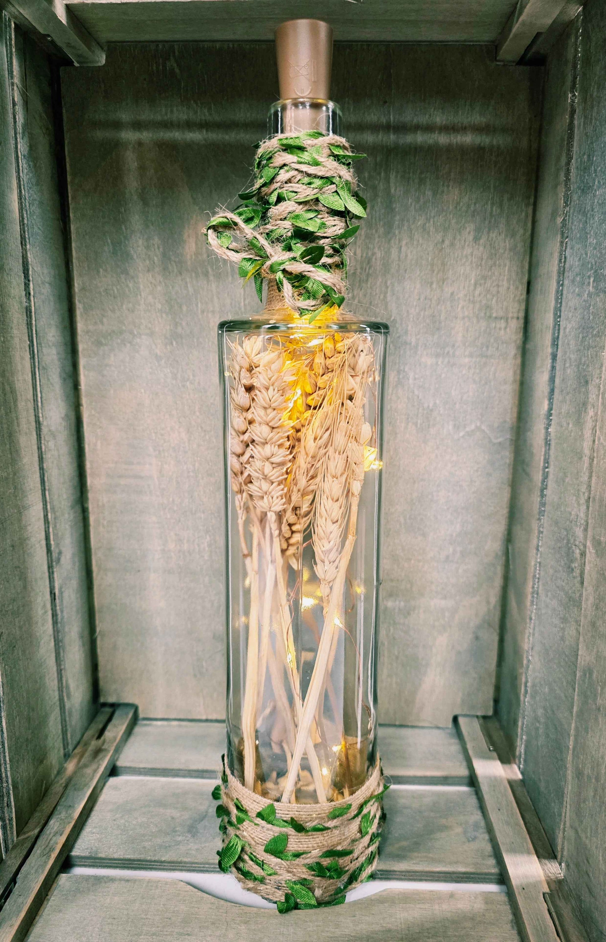 A decorative clear glass bottle with a cork stopper, filled with dried wheat stalks and wrapped with green leaves and natural twine at the neck and base, displayed inside a grey wooden cabinet.