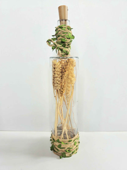 A clear glass bottle with a cork stopper, filled with dried wheat stalks and wrapped with green and beige botanical twine at the neck and base, set against a plain white background.