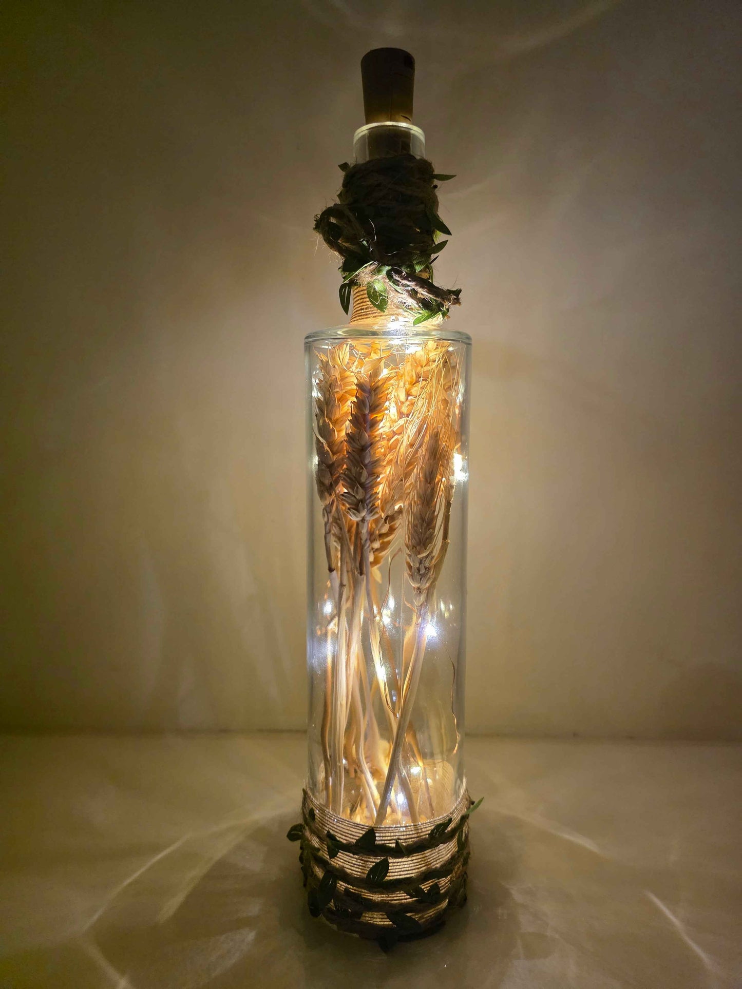 A clear glass bottle with a cork stopper, filled with dried wheat stalks and warm white fairy lights, set against a softly lit beige background.