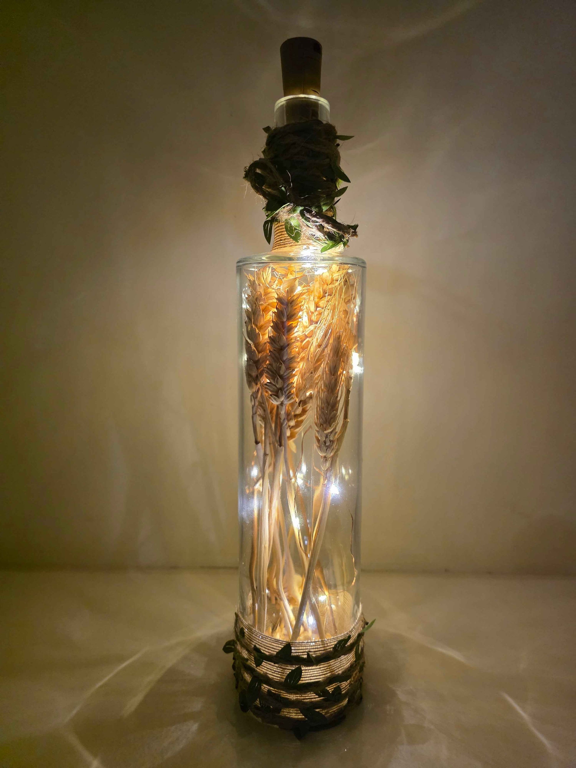 A clear glass bottle with a cork stopper, filled with dried wheat stalks and warm white fairy lights, set against a softly lit beige background.