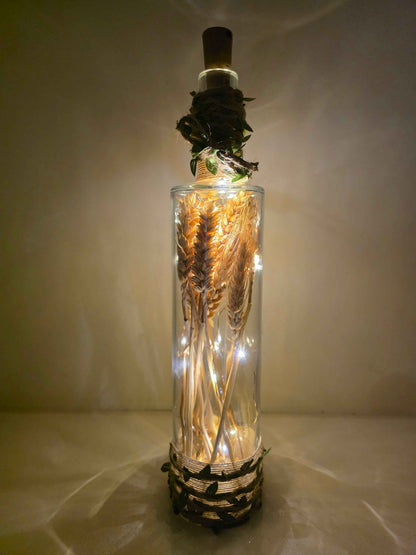 A clear glass bottle with a cork stopper, filled with dried wheat stalks and warm white fairy lights, set against a softly lit beige background.