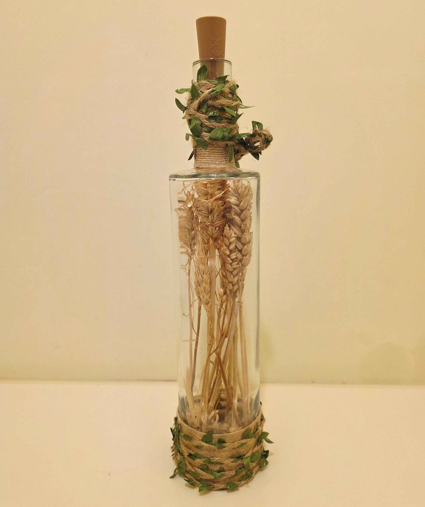 A clear glass bottle with a cork stopper, filled with dried wheat stalks, wrapped with twine and green leaves at the neck and base, set against a plain beige background.