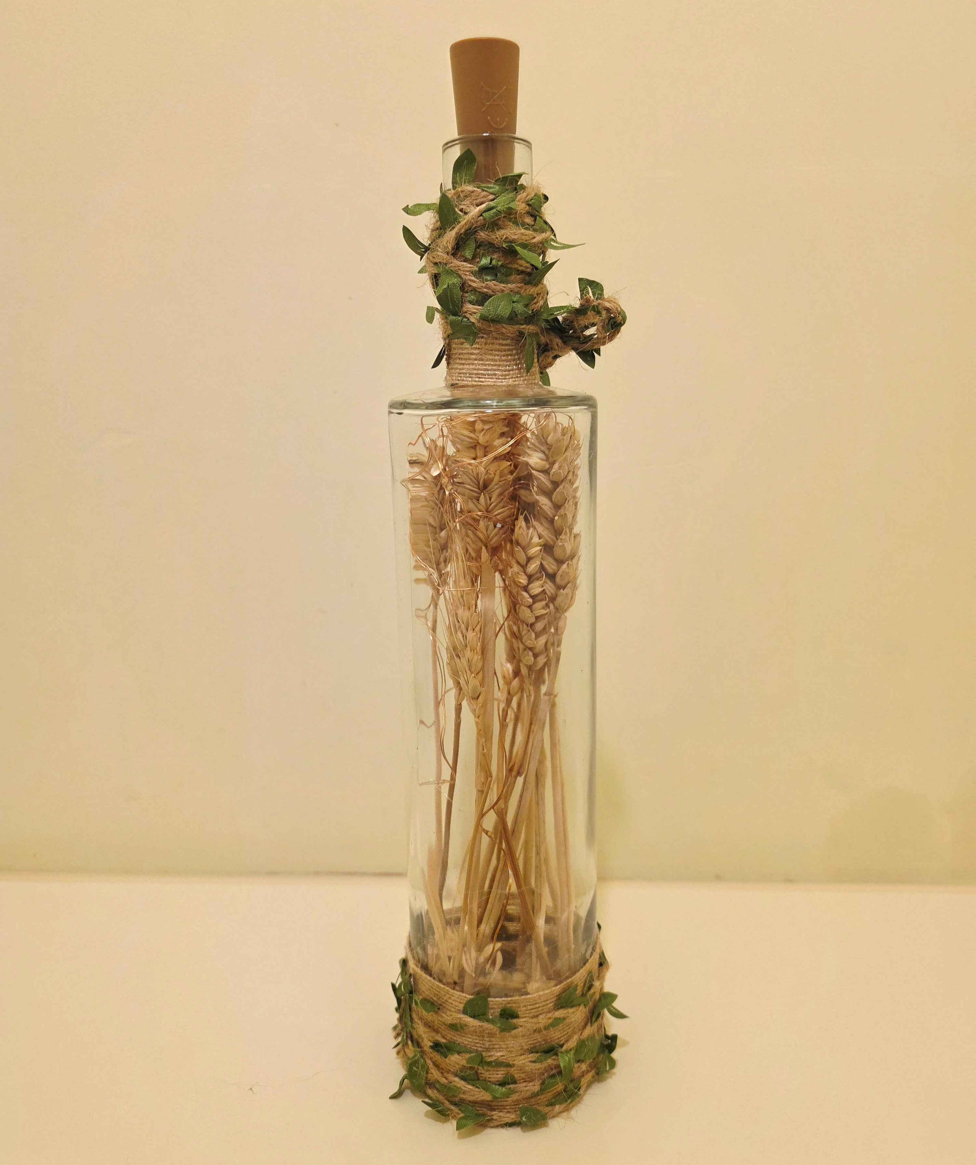 A clear glass bottle with a cork stopper, filled with dried wheat stalks, wrapped with twine and green leaves at the neck and base, set against a plain beige background.