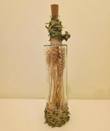 A clear glass bottle with a cork stopper, filled with dried wheat stalks, wrapped with twine and green leaves at the neck and base, set against a plain beige background.