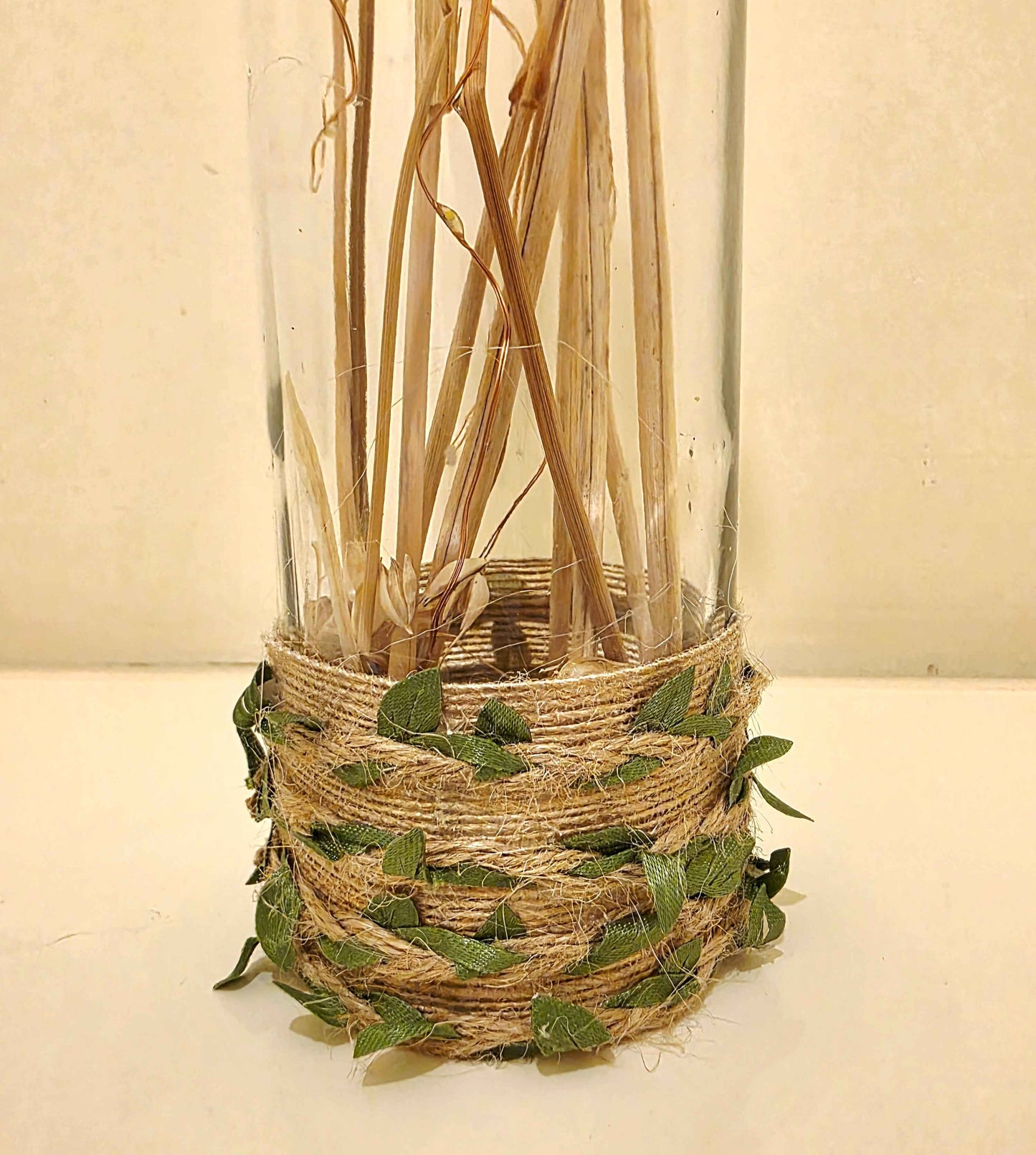 A clear glass bottle filled with dried natural stems, wrapped at the base with tan jute twine and decorated with small green leaves, set against a plain light beige background.