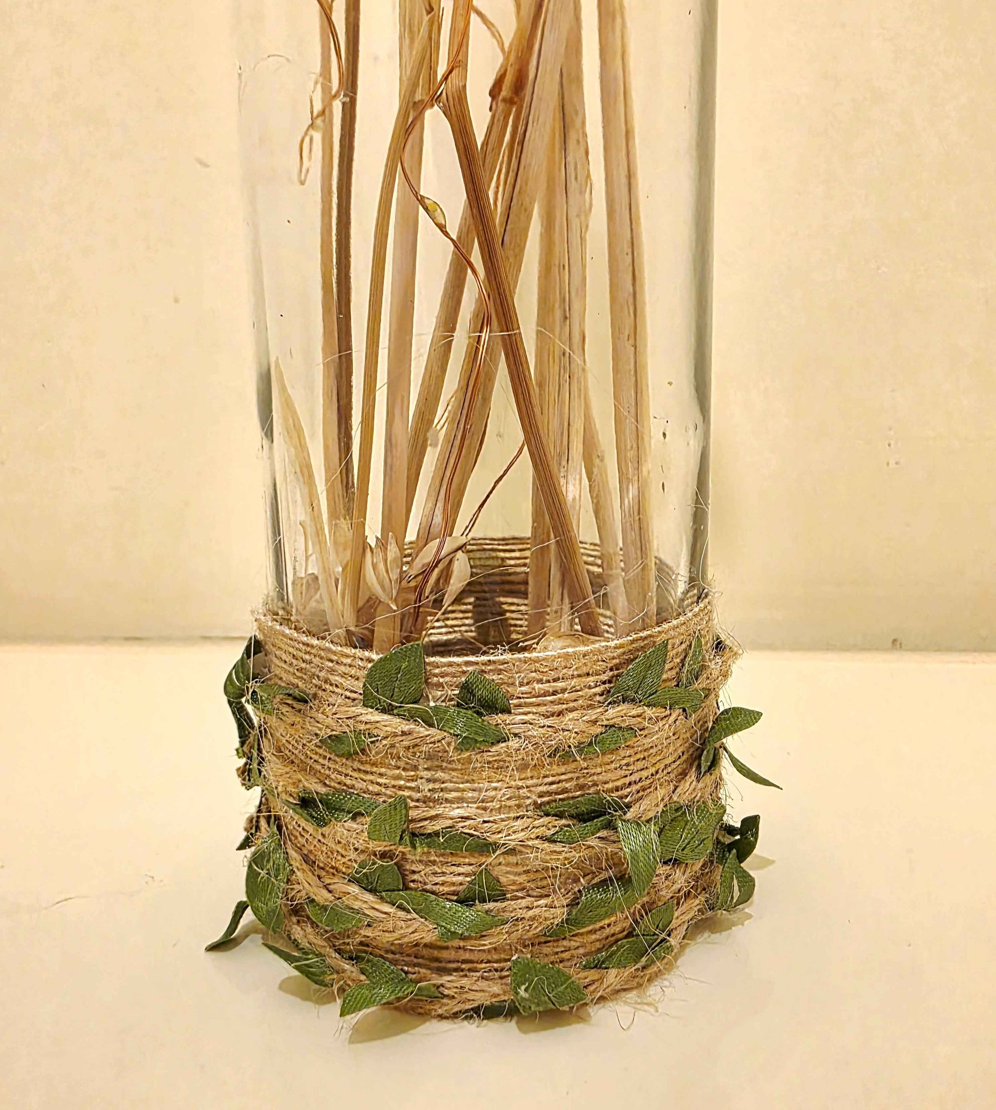 A clear glass bottle filled with dried natural stems, wrapped at the base with tan jute twine and decorated with small green leaves, set against a plain light beige background.
