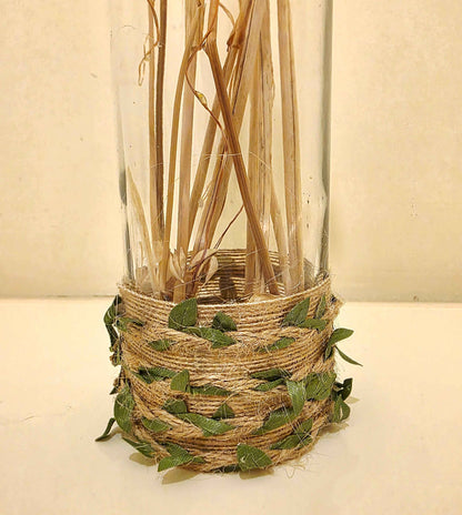 A clear glass bottle filled with dried natural stems, wrapped at the base with tan jute twine and decorated with small green leaves, set against a plain light beige background.
