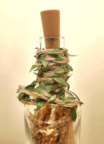 A clear glass bottle filled with dried botanicals and wrapped with natural twine, topped with a brown cork stopper, set against a plain light beige background.