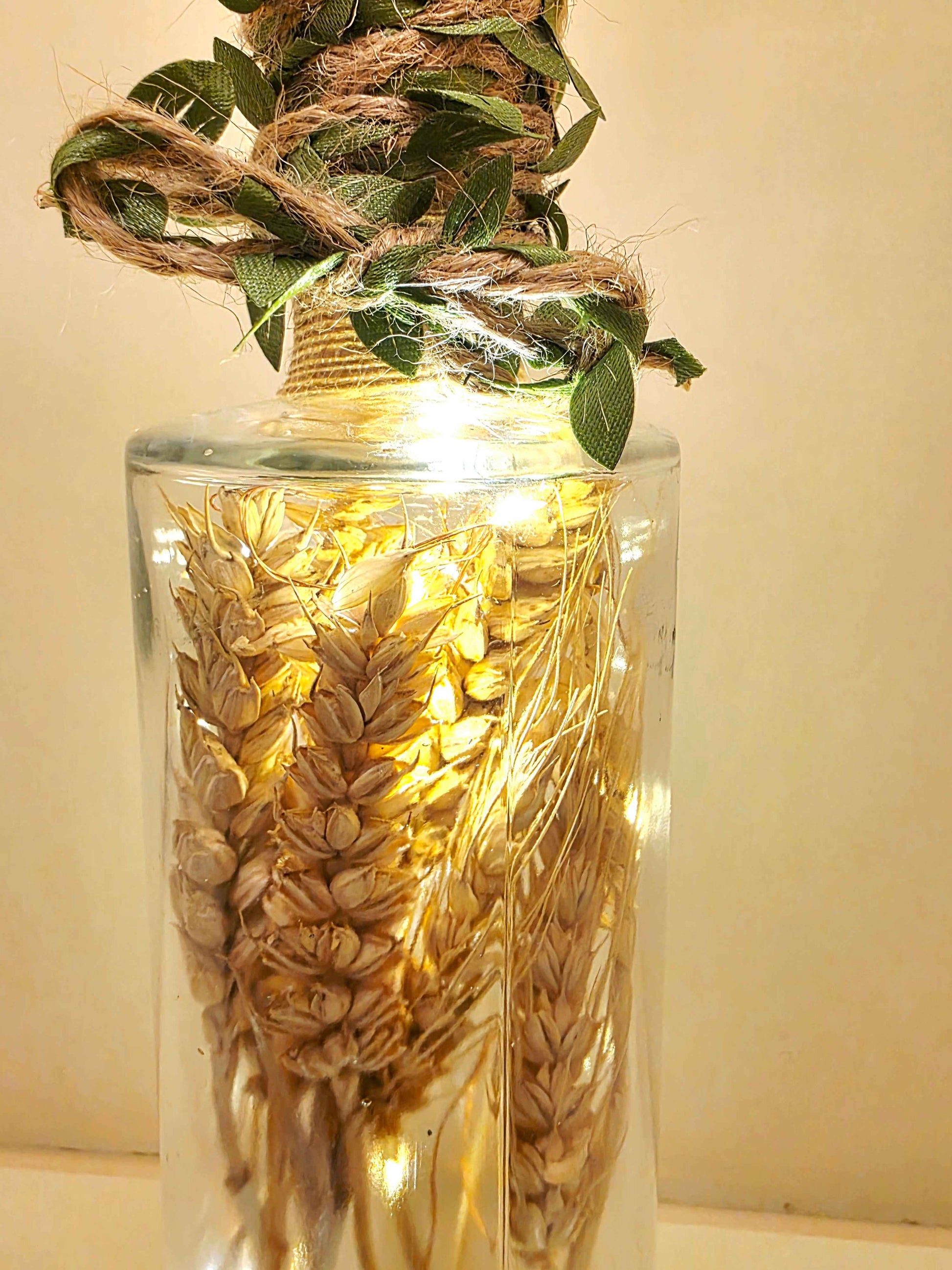 A clear glass bottle filled with dried wheat stalks and wrapped with natural twine and green leaves at the neck, illuminated by warm string lights inside.