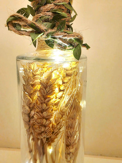 A clear glass bottle filled with dried wheat stalks and wrapped with natural twine and green leaves at the neck, illuminated by warm string lights inside.