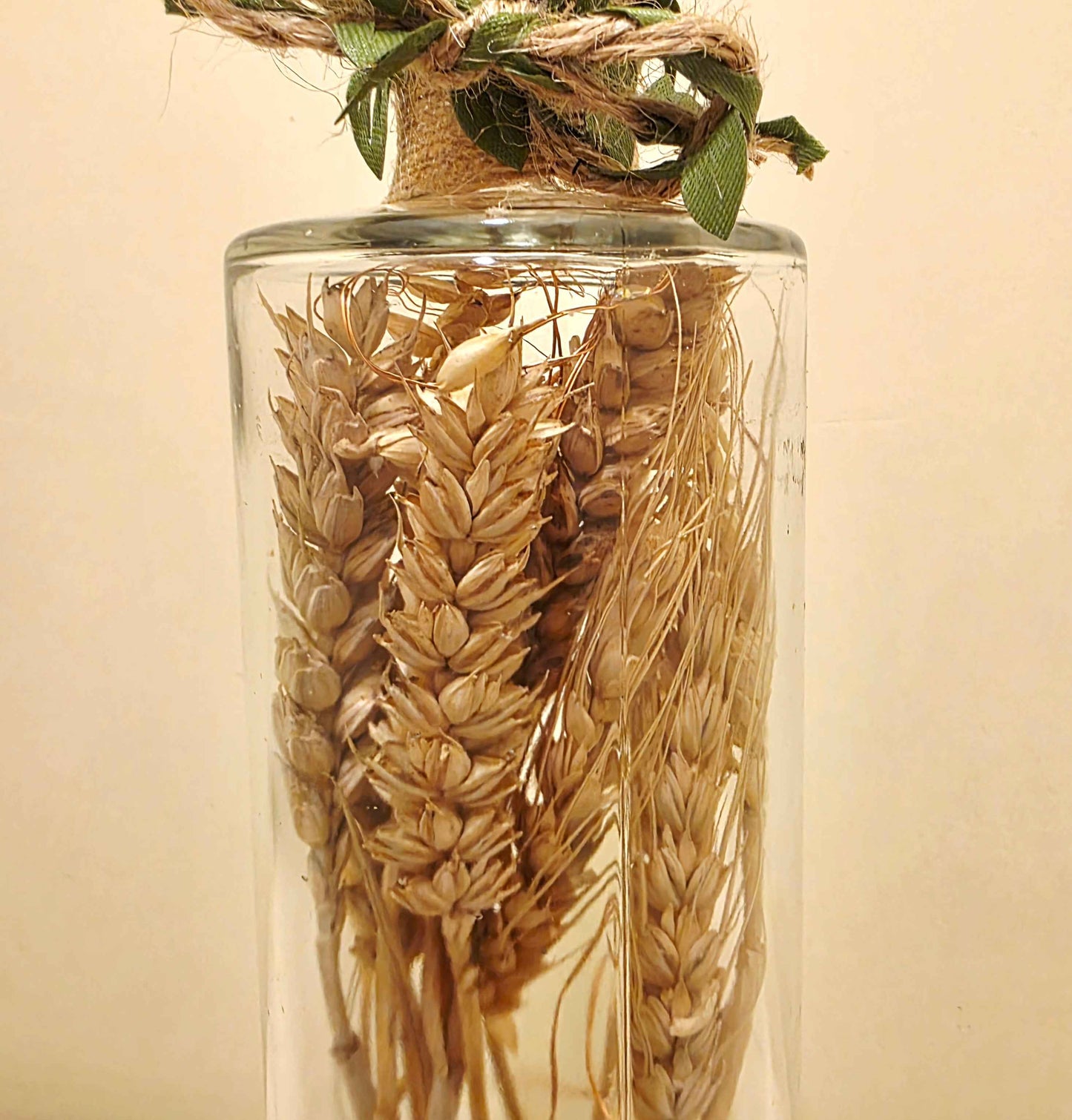 A clear glass bottle filled with dried wheat stalks, tied at the neck with twine and green leaves, against a plain beige background.