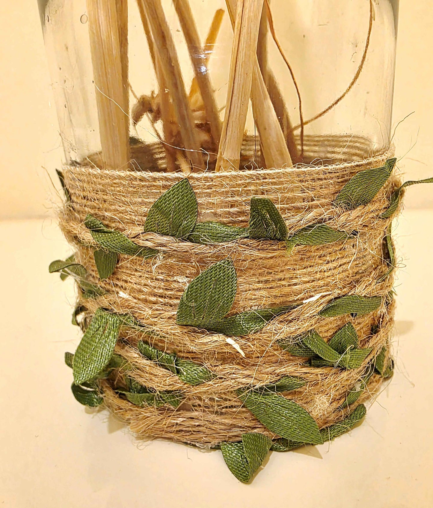 A decorative clear glass jar wrapped with natural jute twine and green fabric leaves, filled with dried reeds or stems, photographed against a plain light background.