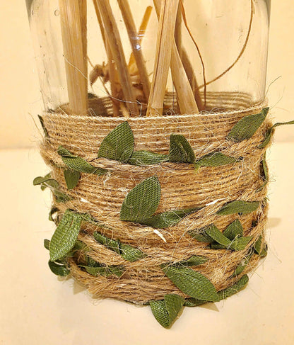 A decorative clear glass jar wrapped with natural jute twine and green fabric leaves, filled with dried reeds or stems, photographed against a plain light background.