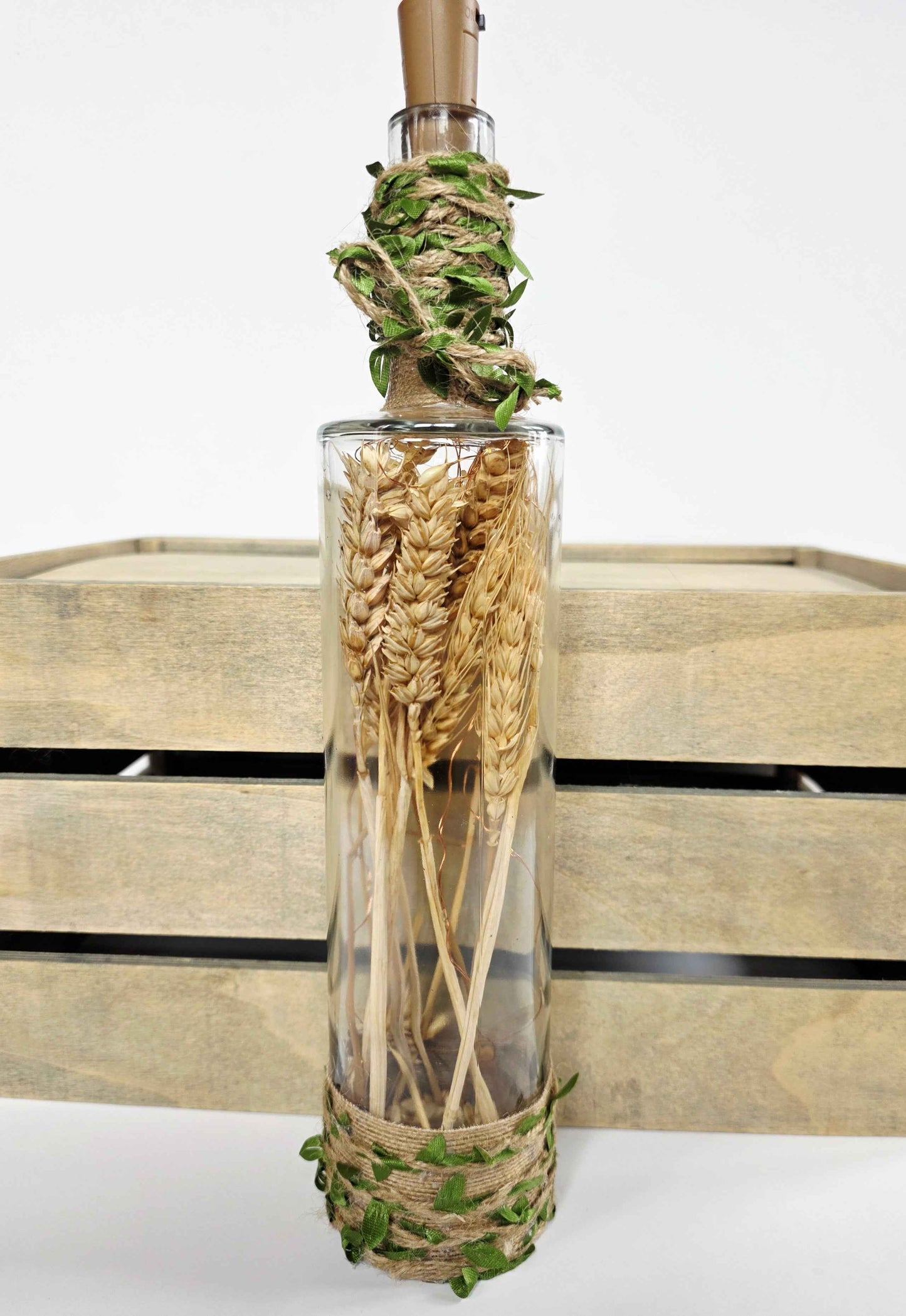 A clear glass bottle with a cork stopper, filled with dried wheat stalks, wrapped with twine and green leaves at the top and bottom, set against a rustic wooden crate background.