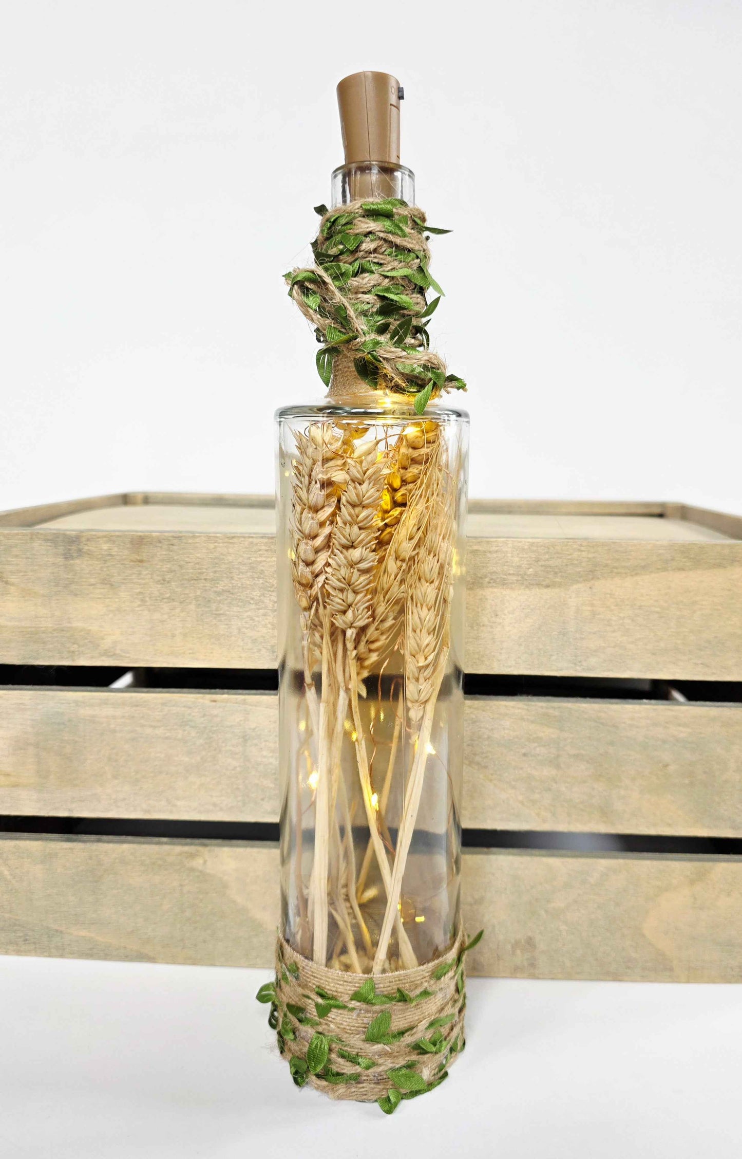 A clear glass bottle filled with dried wheat stalks, wrapped with green vines at the neck and base, and topped with a cork stopper, displayed in front of a rustic wooden crate on a white background.