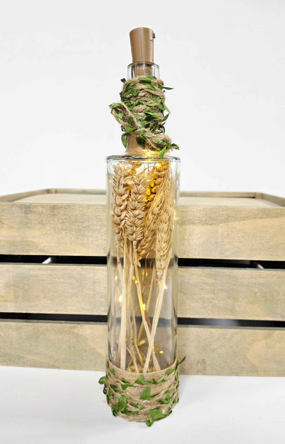A clear glass bottle filled with dried wheat stalks, wrapped with green vines at the neck and base, and topped with a cork stopper, displayed in front of a rustic wooden crate on a white background.