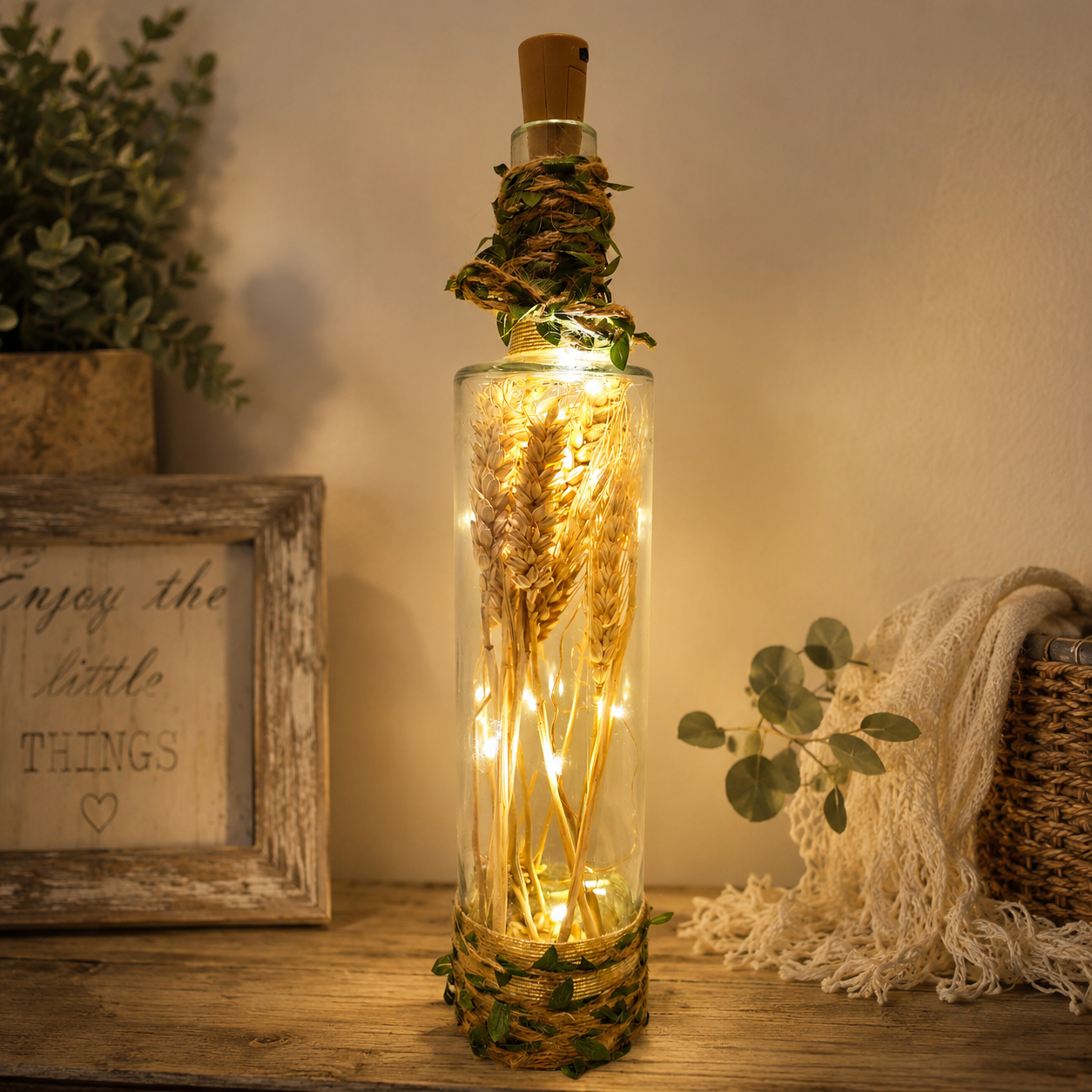 A clear glass bottle with a cork stopper, wrapped in twine and greenery, filled with dried wheat stems and warm white fairy lights, displayed on a wooden surface in a cozy home setting.
