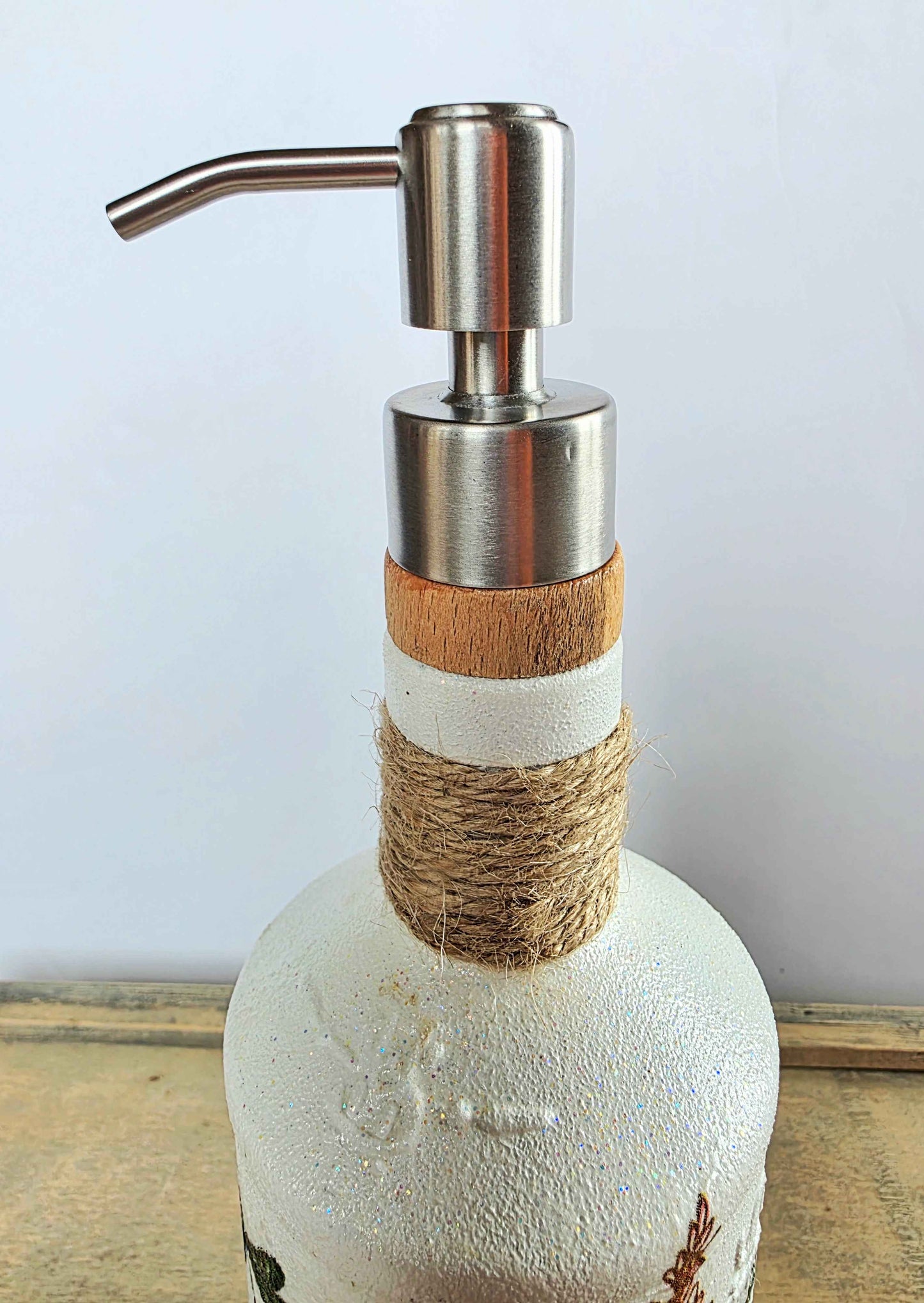 A rustic soap dispenser made from a white glass bottle, wrapped with natural jute twine and a light wood band, topped with a brushed stainless steel pump dispenser.