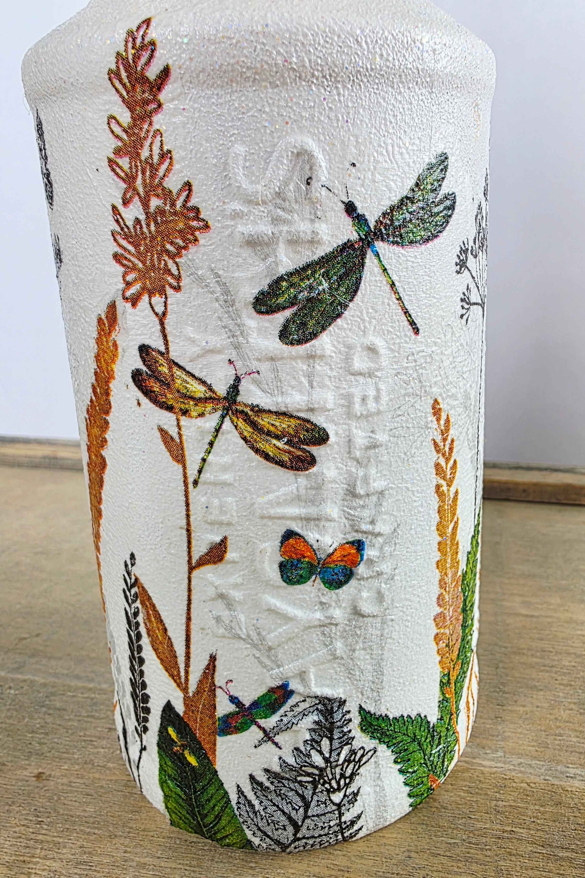 A white textured glass bottle featuring a detailed illustration of dragonflies, butterflies, leaves, and dried grasses