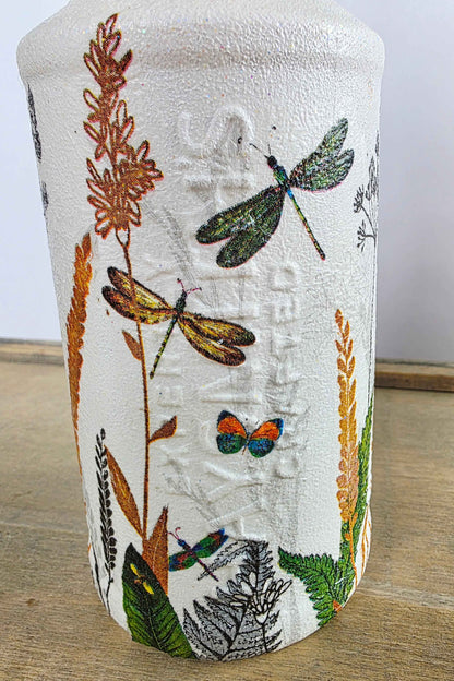 A white textured glass bottle featuring a detailed illustration of dragonflies, butterflies, leaves, and dried grasses