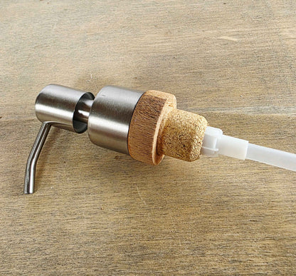 A close-up of a pump dispenser with a brushed stainless steel body, a natural cork accent ring, and a white plastic tube, resting on a light brown wooden surface.