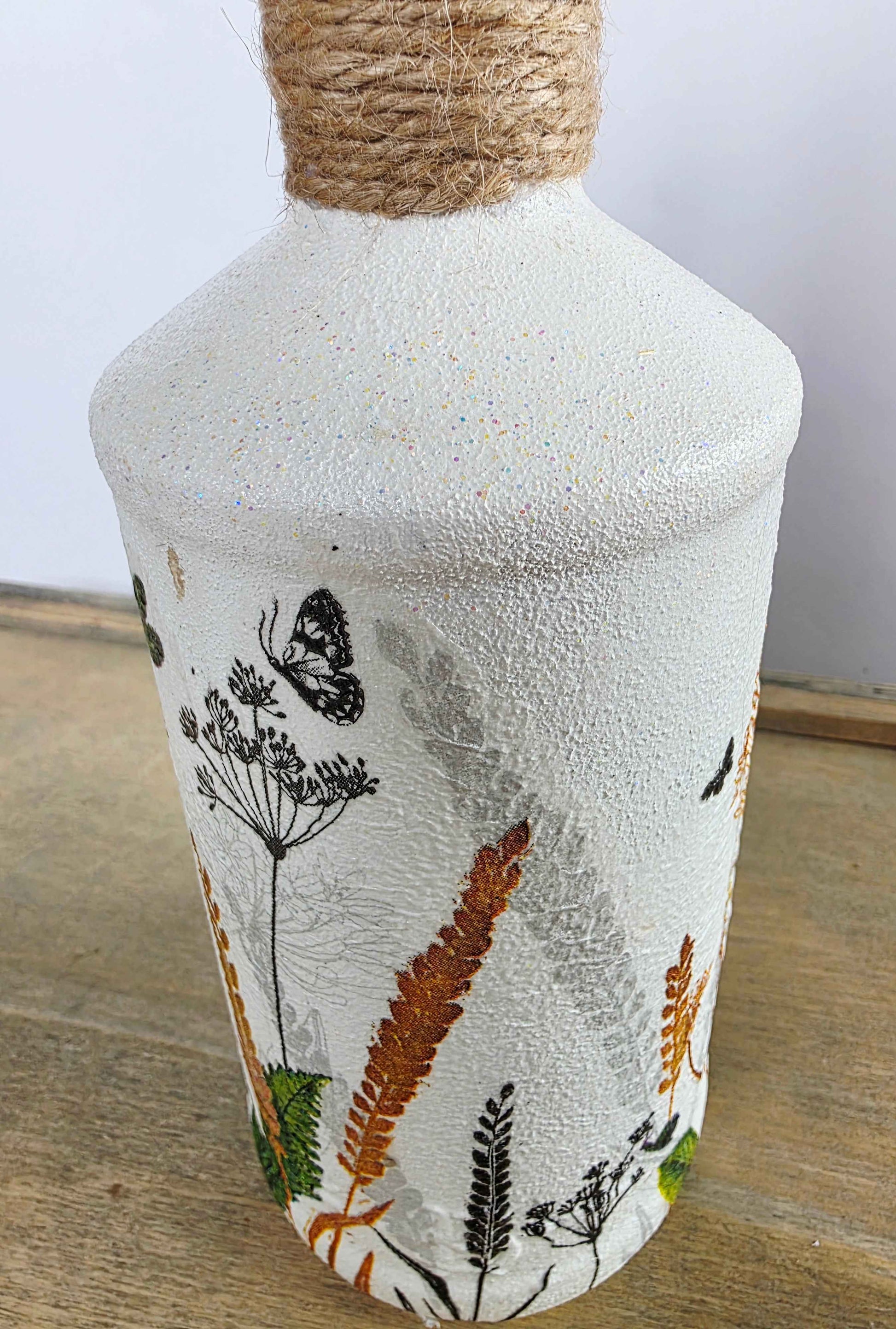 A textured off-white glass bottle with a natural jute rope-wrapped neck, decorated with botanical illustrations of ferns, wildflowers, and butterflies in black, orange, and green, resting on a wooden surface.