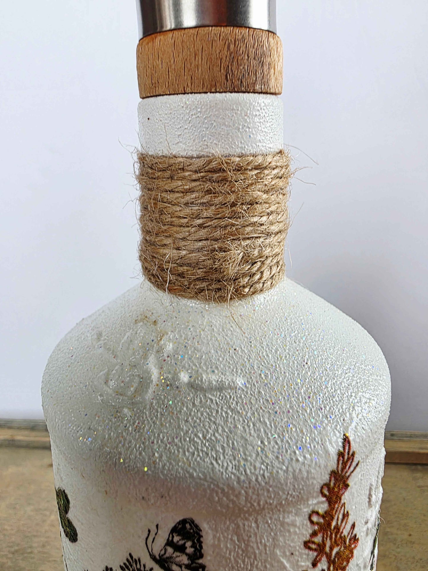 A close-up of a white textured bottle with a natural wood stopper, wrapped in brown twine at the neck, and decorated with black and brown botanical illustrations.