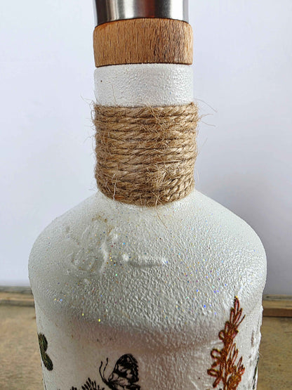 A close-up of a white textured bottle with a natural wood stopper, wrapped in brown twine at the neck, and decorated with black and brown botanical illustrations.