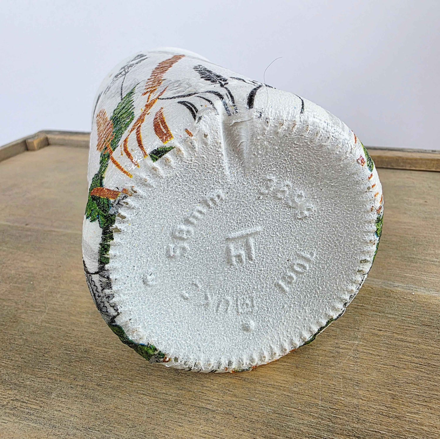A white painted bottle with a colourful floral pattern around the exterior, shown upside down on a wooden surface.