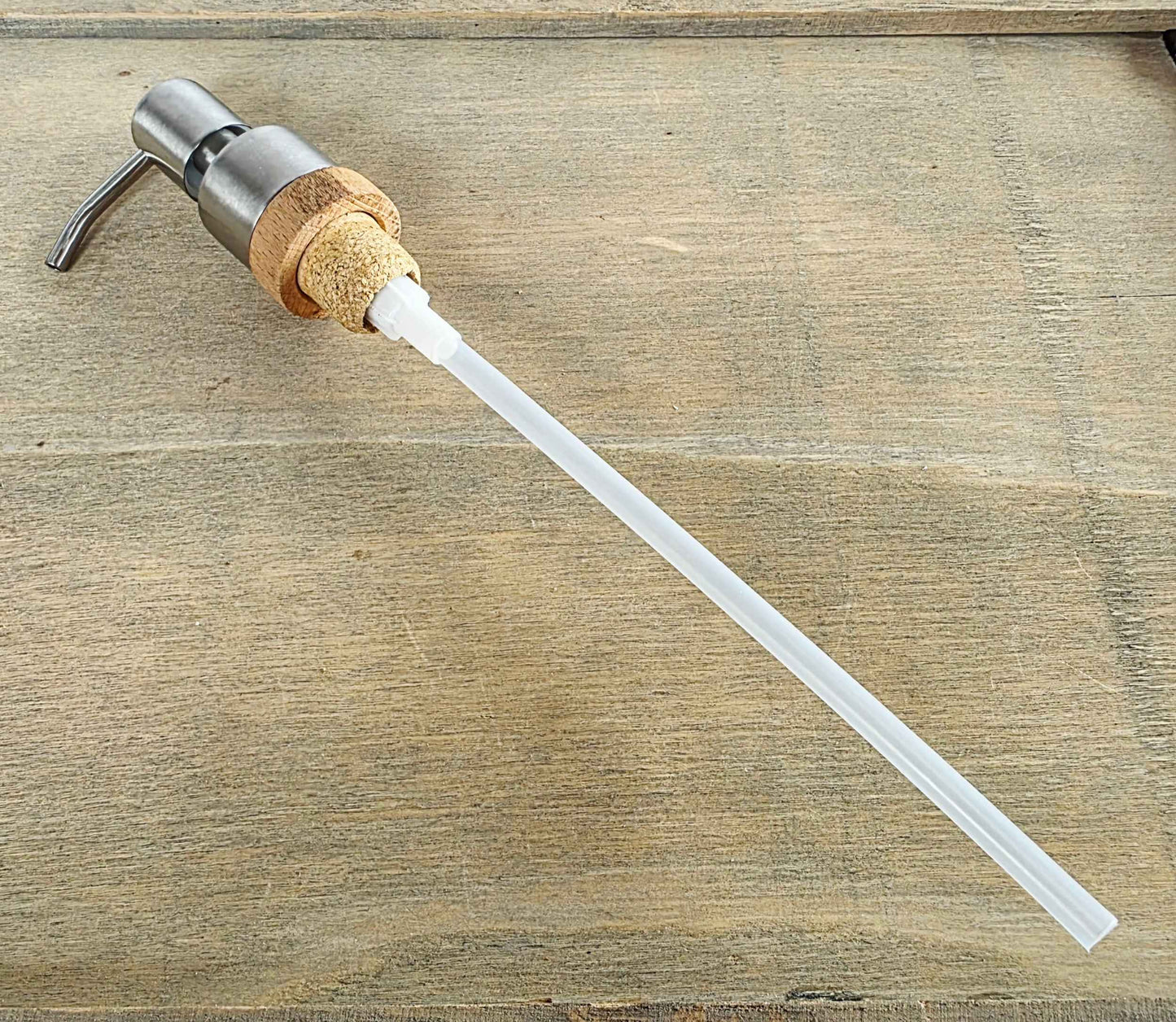 A pump dispenser with a brushed metal pump head, a natural cork collar, and a long translucent white tube, shown on a rustic wood surface.