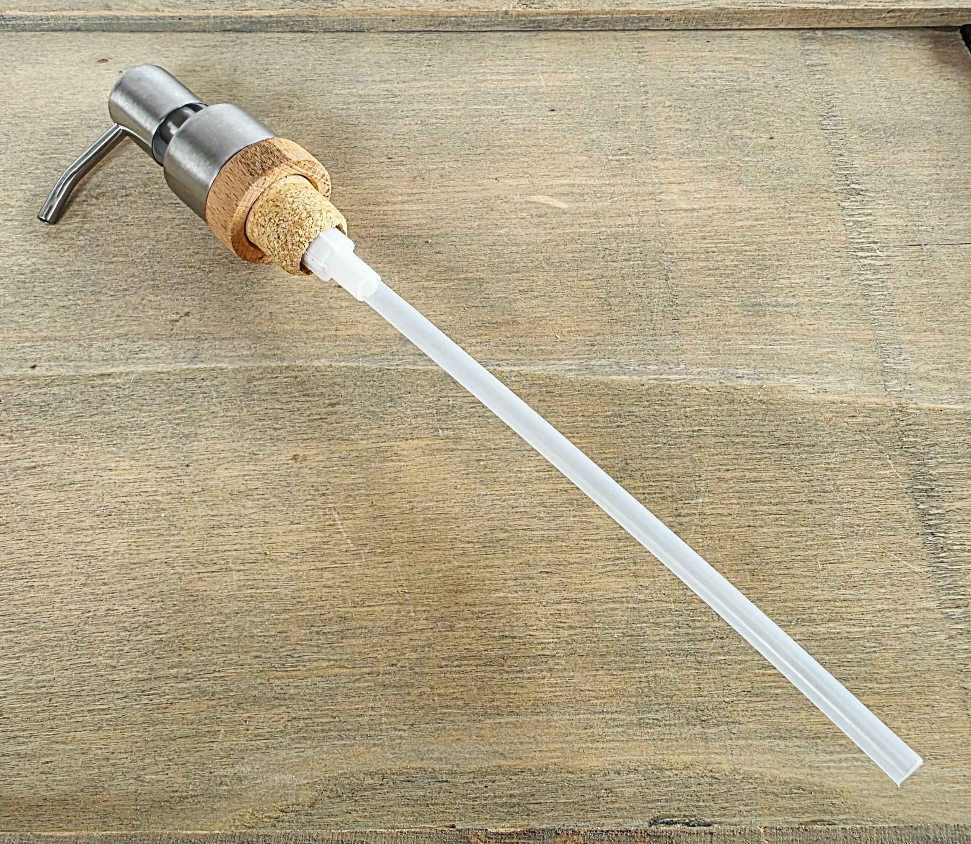 A pump dispenser with a brushed metal pump head, a natural cork collar, and a long translucent white tube, shown on a rustic wood surface.