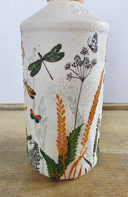A handmade white painted bottle with a textured off-white glaze, decorated with hand-painted botanical and insect motifs including dragonflies, butterflies, and wildflowers in green, orange, and black.