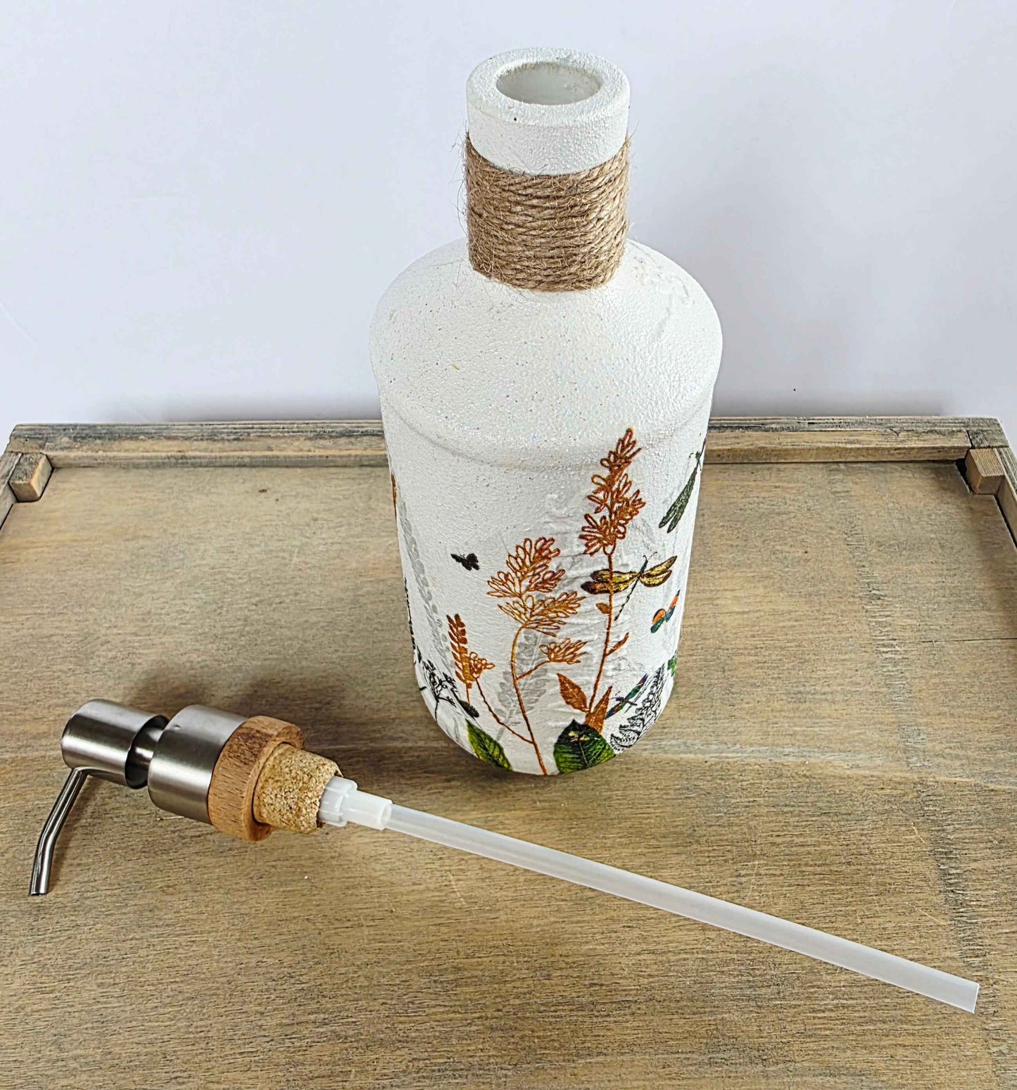 A decorative white glass bottle with botanical illustrations and a wrapped neck, shown on a wooden tray with a metal-and-cork pump dispenser and clear transfer tube in front.