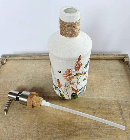 A decorative white glass bottle with botanical illustrations and a wrapped neck, shown on a wooden tray with a metal-and-cork pump dispenser and clear transfer tube in front.
