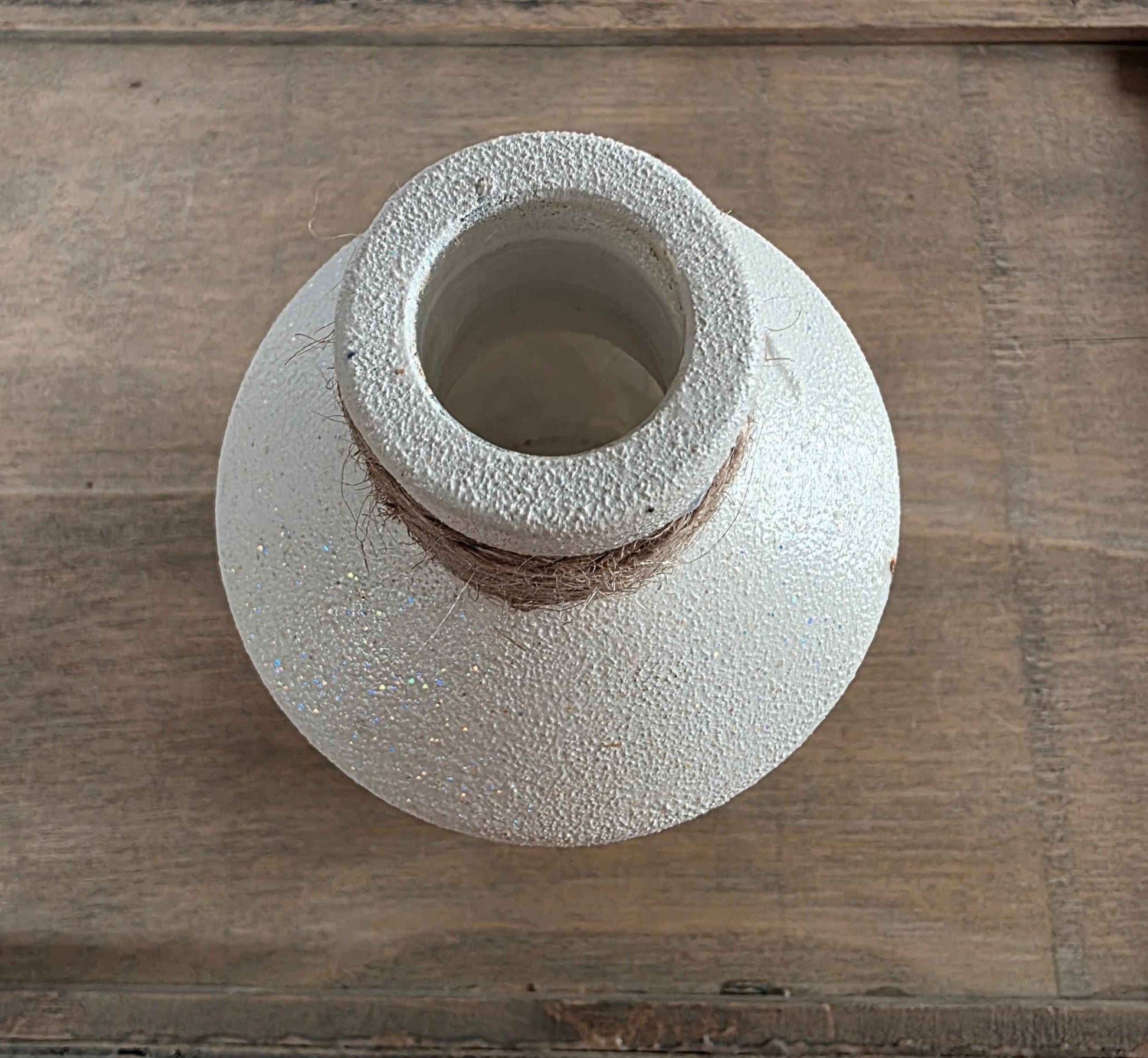 A handmade glass bottle with a textured, speckled off-white glaze and a rustic brown clay band around the neck, shown on a wooden surface.